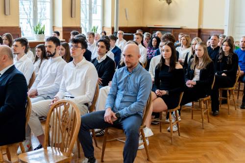 Młodzież galowo ubrana siedzi na jasnych krzesłach