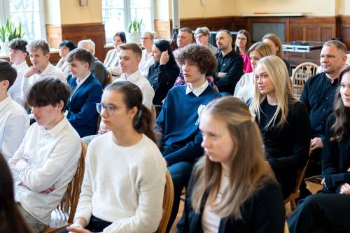 Młodzież galowo ubrana siedzi na jasnych krzesłach