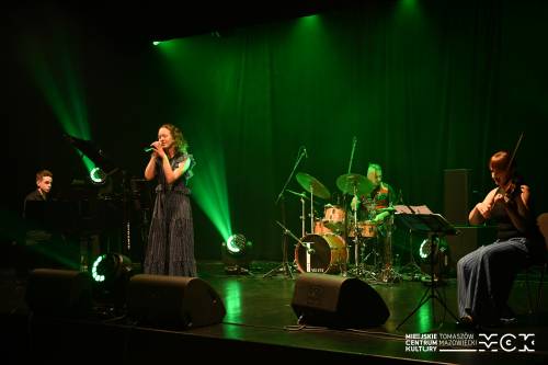 Na zdjęciu koncert zespołu Arete z wokalistką przy mikrofonie. Kolorowe światła sceniczne. Na drugim planie pozostali muzycy